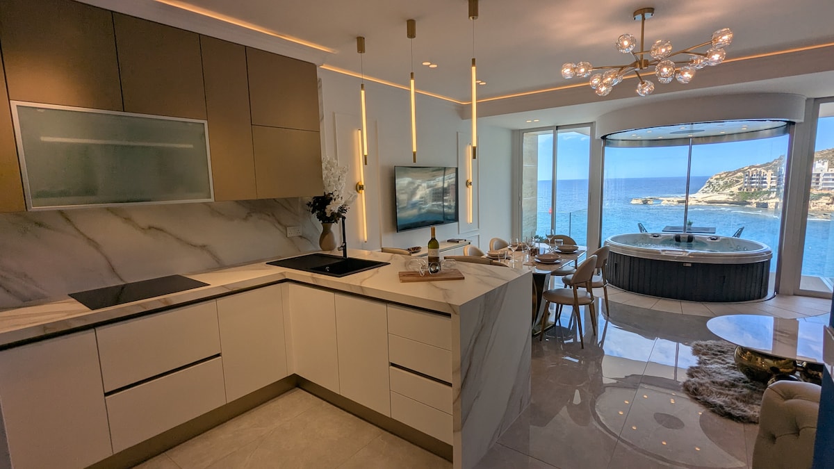 A modern kitchen is highlighted by sleek cabinetry and a marble countertop, featuring a central island with a cooktop. Large windows provide an expansive view of the sea, while a circular spa bath is visible in the adjacent area, enhancing the coastal ambiance.