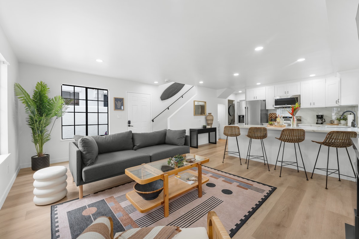 A modern living space features a spacious gray sofa, accompanied by wood-framed bar stools at a sleek kitchen island. Light wood flooring extends throughout, while a patterned area rug adds texture. Natural light enters through large windows, enhancing the bright and open atmosphere.