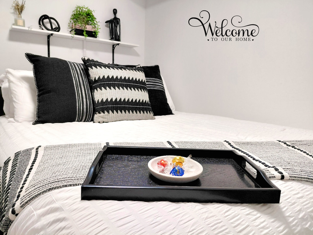 A cozy queen-size bed is adorned with black and white patterned pillows and decorative throws. A tray with treats is placed on the bed, adding a welcoming touch. A wall-mounted shelf features a small plant and decorative statue, while a 'Welcome to Our Home' sign decorates the wall.