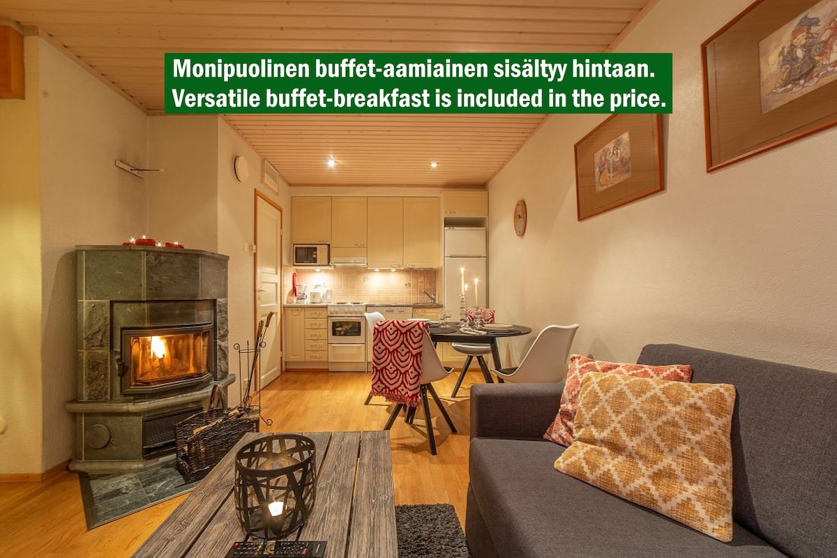 A cozy living area features a comfortable sofa and a dining table for four. A wood-burning stove adds warmth to the space. The kitchenette, equipped with modern appliances, is located in the background, complemented by soft lighting and wooden paneling.