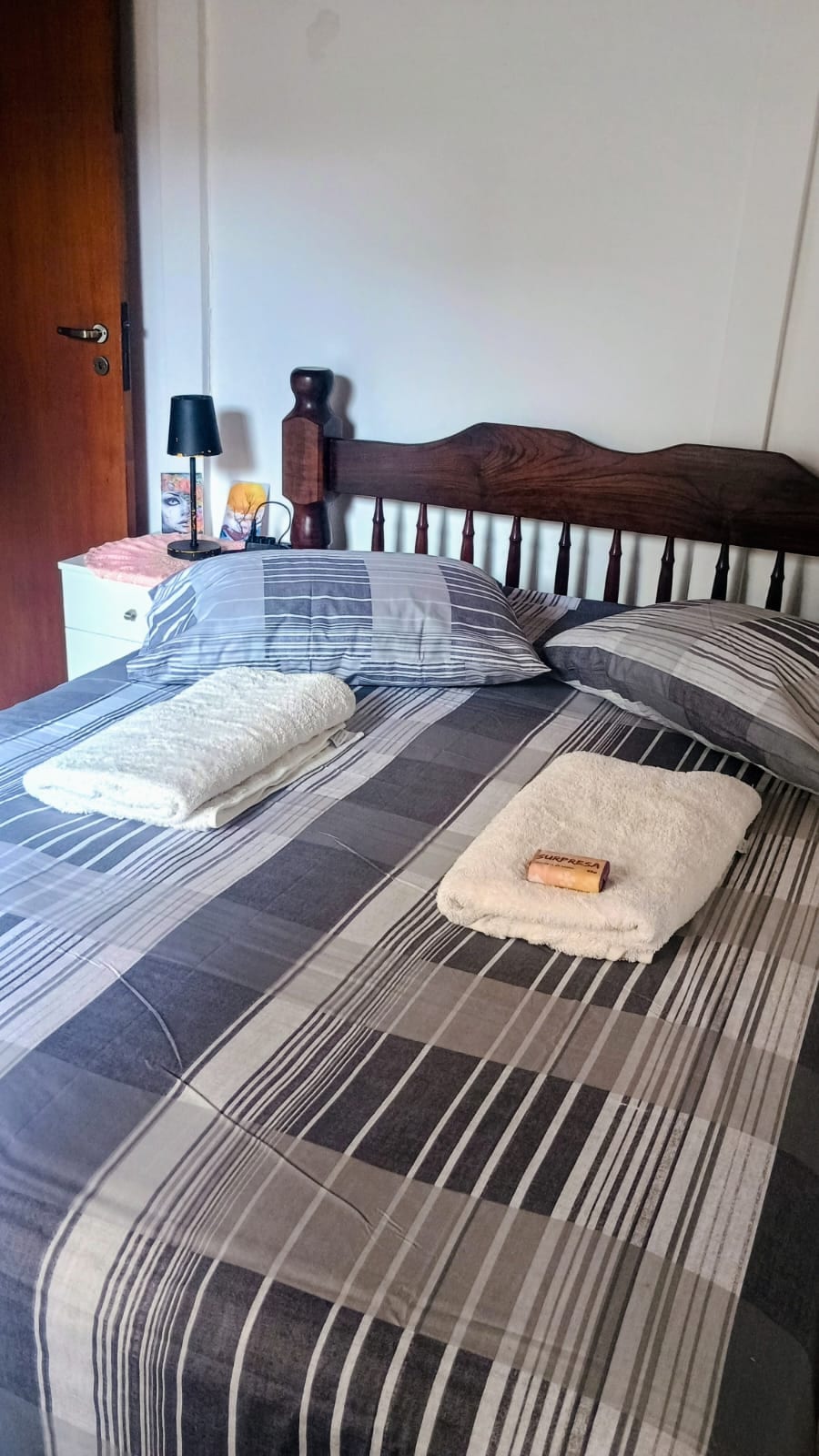 A spacious bed is dressed in a striped bedding set, featuring neutral colors. Two rolled towels are placed neatly on top. A bedside table with a lamp and personal items is positioned beside the bed. Natural light enters through a nearby door.