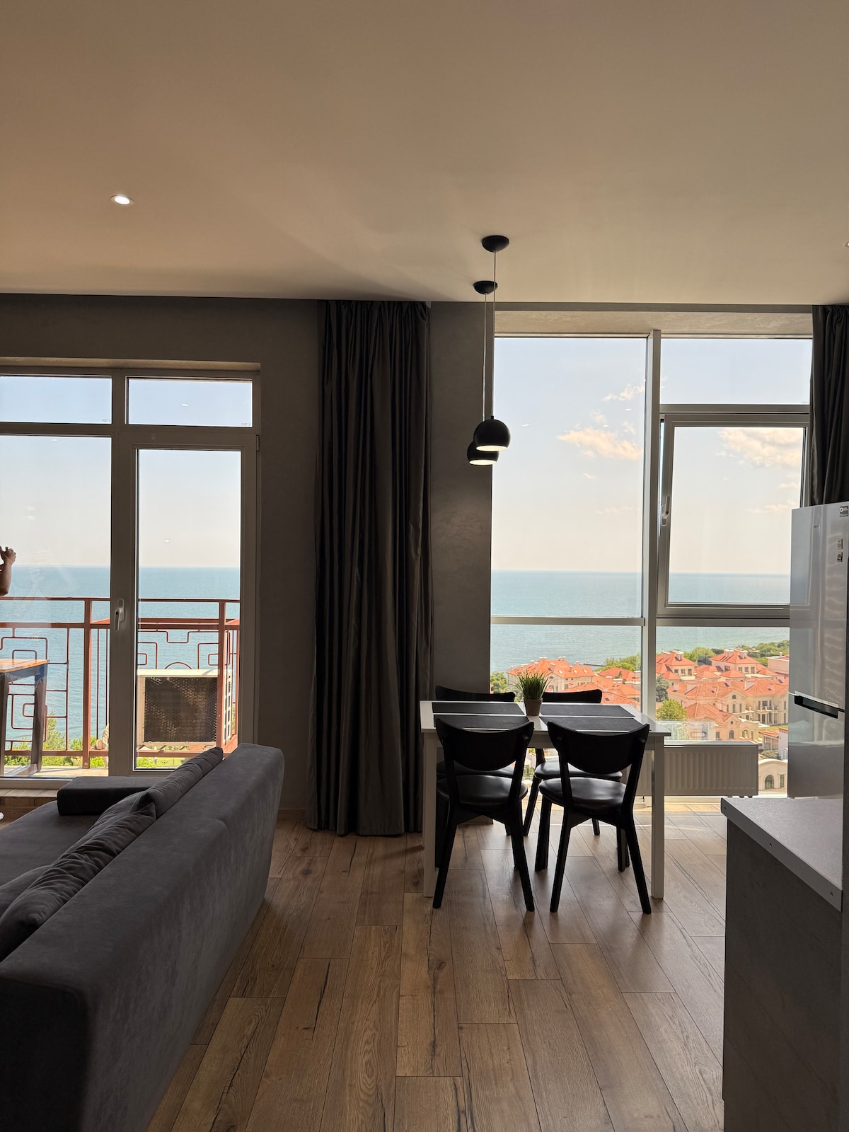 A modern living space features a sleek sofa near large windows offering views of the sea. A dining table for four is positioned by the window, with natural light illuminating the interiors. Light-colored walls and wooden flooring enhance the overall ambiance.