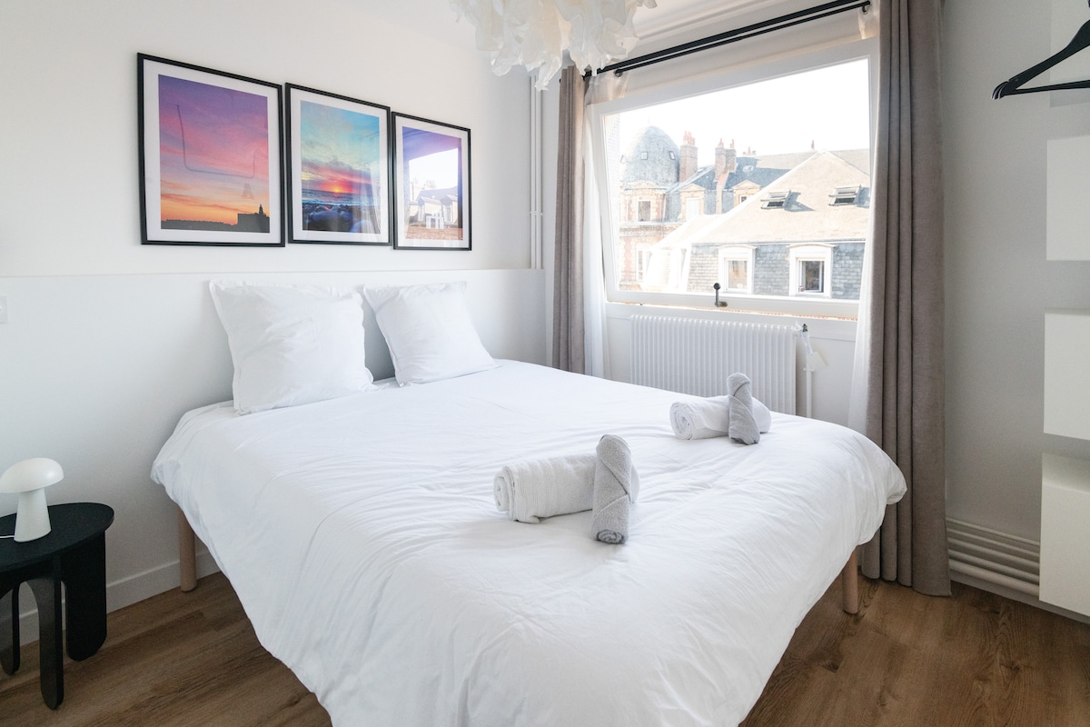 A spacious sleeping area is presented with a large bed featuring crisp white linens. Above the bed, three framed artworks depict colorful landscapes. Natural light floods the room from a window overlooking the rooftops, complemented by sheer curtains that softly filter the sunlight.
