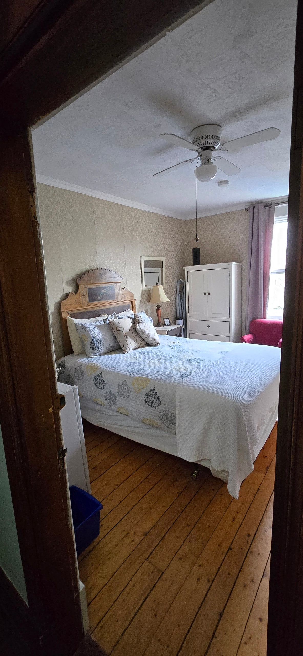 A doorway frames a cozy room featuring a queen-size bed adorned with decorative pillows. Natural light filters through a window dressed with curtains, illuminating hardwood floors. A dresser and small chair add functional elements, while a ceiling fan provides comfort.