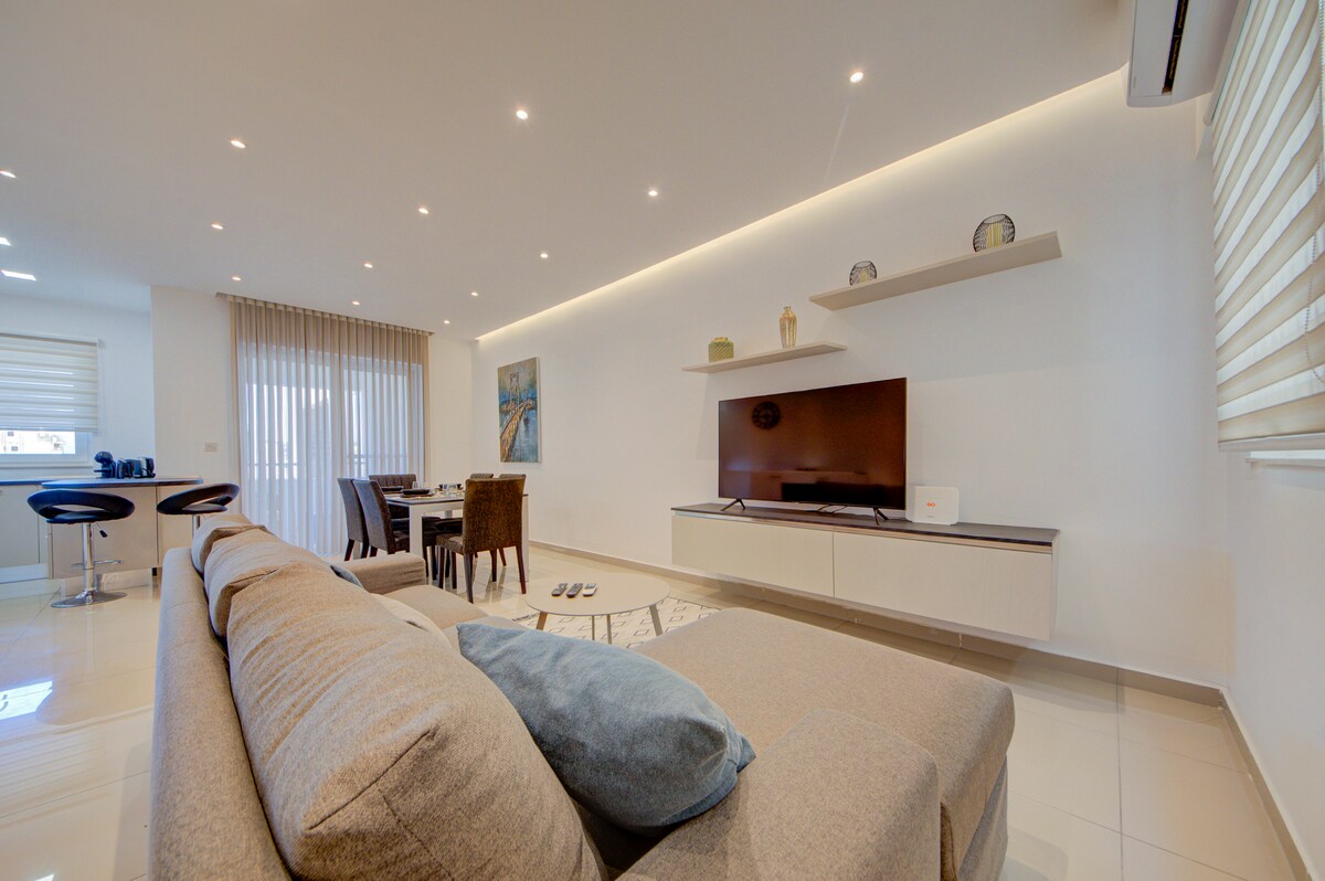 An open-plan living area features a spacious sectional sofa in neutral tones accented by a blue cushion. A flat-screen TV is mounted on a white wall unit, while a dining set and bar stools create a welcoming atmosphere. Natural light filters through the window treatments.