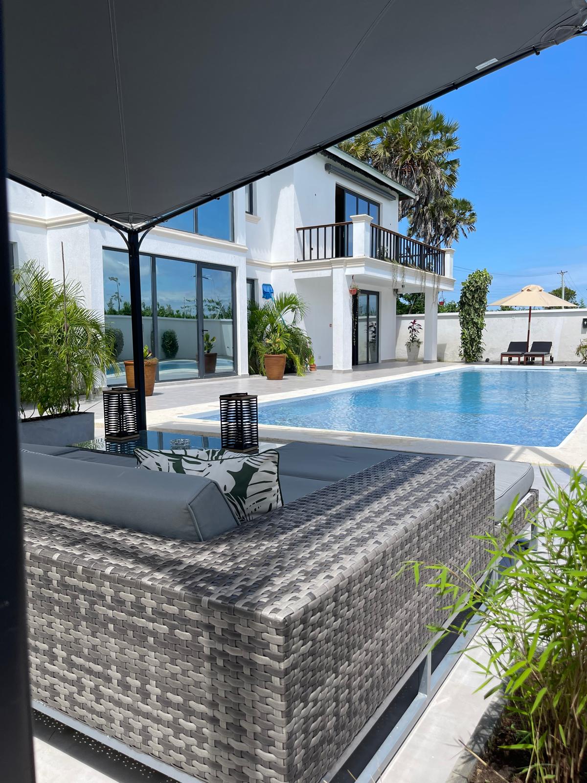 A stylish outdoor seating area features a comfortable couch with patterned cushions, positioned near a shimmering pool surrounded by lush greenery. Large windows of the apartment reflect the bright blue sky, creating a serene atmosphere perfect for relaxation.
