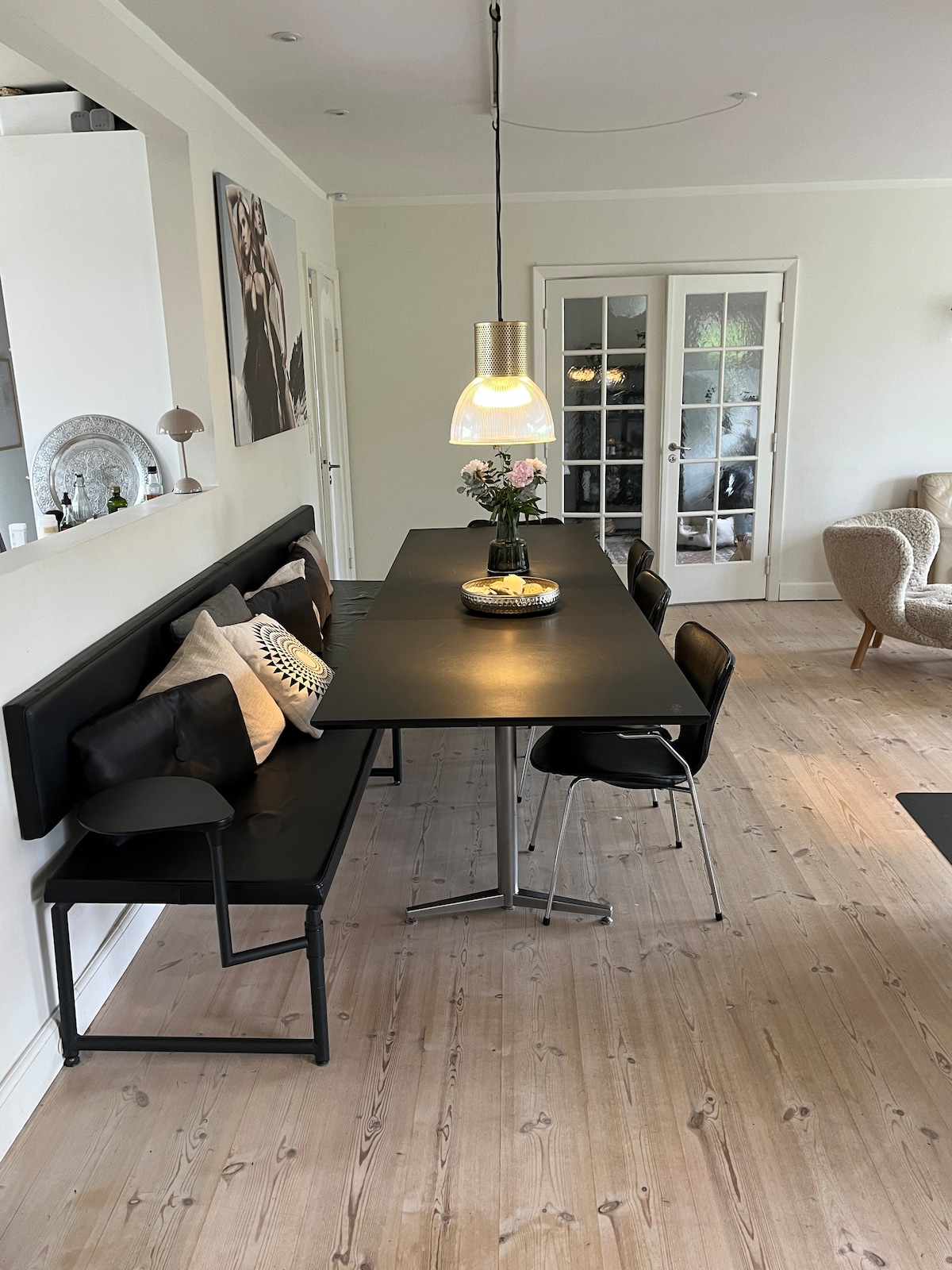 A modern dining area features a sleek black table surrounded by a mix of chairs and a bench. A pendant light hangs above the table, creating focused illumination. Decorative pillows are placed on the bench, adding a touch of comfort to the space.