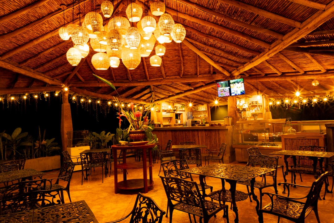 The dining area features an open structure with a thatched roof supported by wooden beams. Ornate metal tables and chairs are arranged throughout, and a cluster of decorative light fixtures hangs overhead, casting a warm glow. A bar area is visible in the background, enhancing the inviting space.