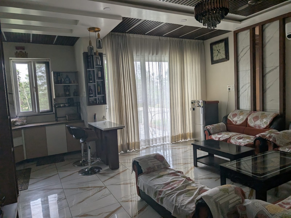 A bright living area features large windows adorned with sheer curtains, allowing natural light to fill the space. Comfortable seating is arranged around a glass coffee table, and a stylish bar counter is visible. The room is decorated with modern accents and a wall clock.