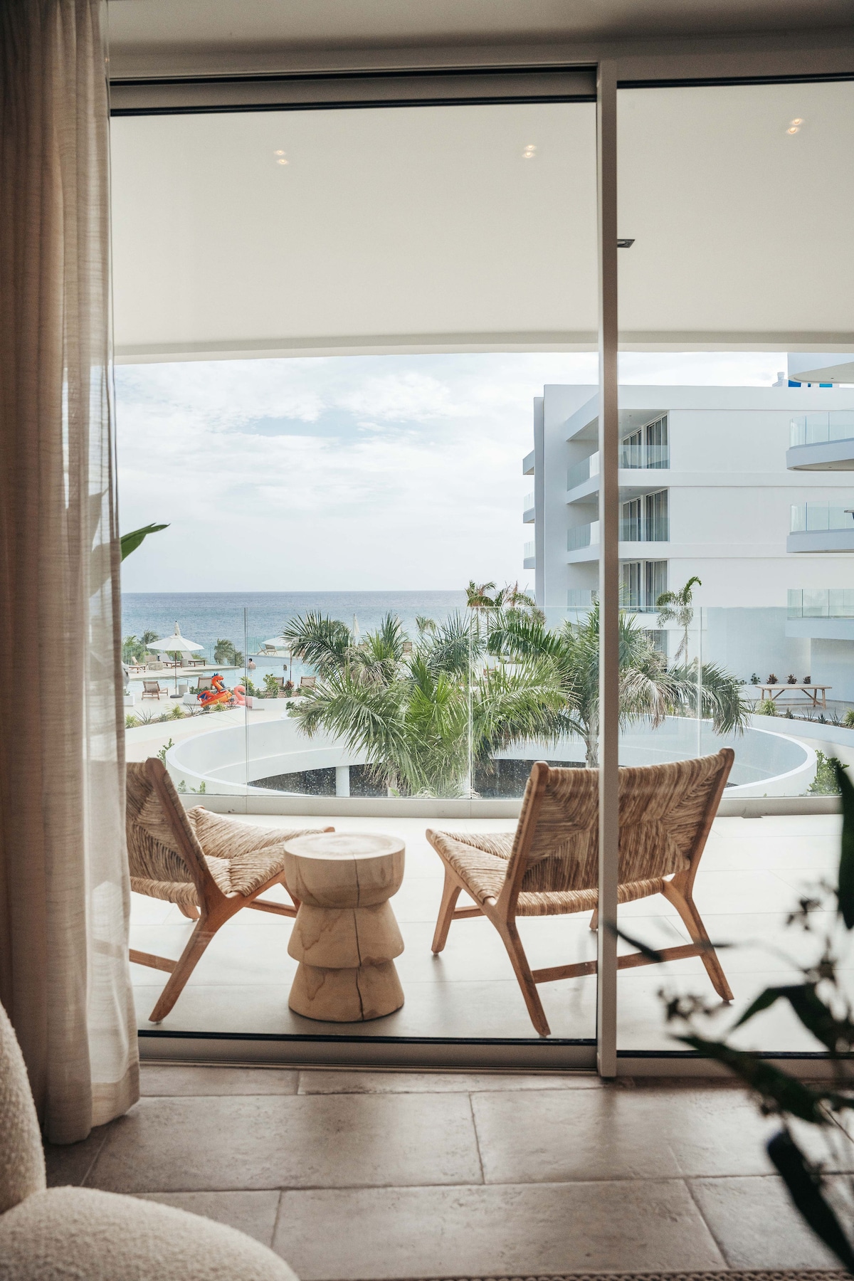 A spacious balcony area is visible through large glass doors, featuring two woven chairs and a small wooden side table. Lush greenery and the ocean can be seen in the background, suggesting a serene outdoor space.