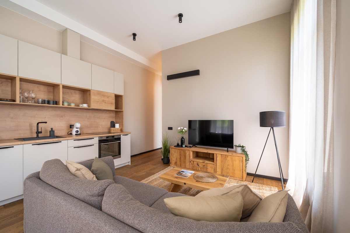 A modern living area features a comfortable, spacious sofa paired with accent pillows. A wooden coffee table sits in front, and a sleek television is positioned on a light wooden media console. A floor lamp adds soft lighting, and natural light enters through large windows with sheer curtains.