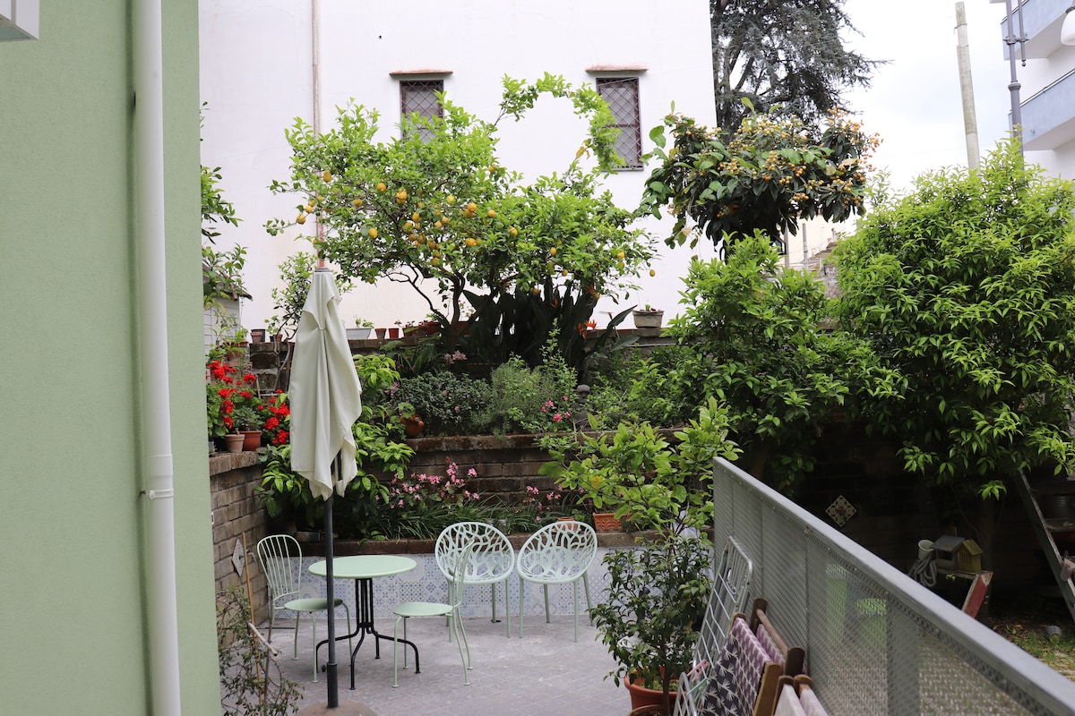 A shared outdoor patio features a round table and several light green chairs surrounded by a lush garden. Various plants and vibrant flowers create a serene environment, highlighted by a large tree bearing citrus fruits. The space is designed for relaxation and enjoyment of nature.