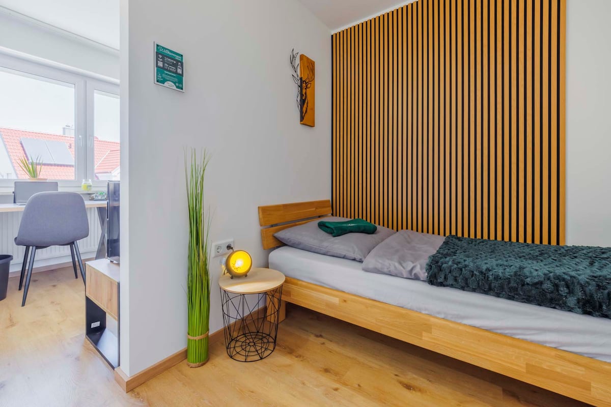 A minimalist bedroom features a single bed with a wooden frame and soft bedding. A striped wooden accent wall adds character. A small round table with a lamp sits beside the bed. A workspace with a chair is visible in the background, along with a large window allowing natural light.