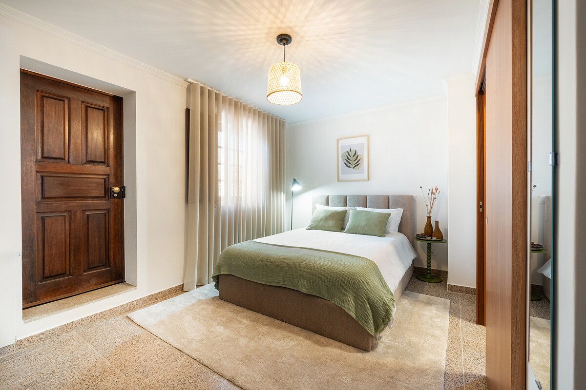 A serene bedroom features a spacious bed with soft green accents and neutral bedding. Natural light filters through a window adorned with light drapes. A decorative plant and artwork add subtle touches, while a wooden door provides access to the room.