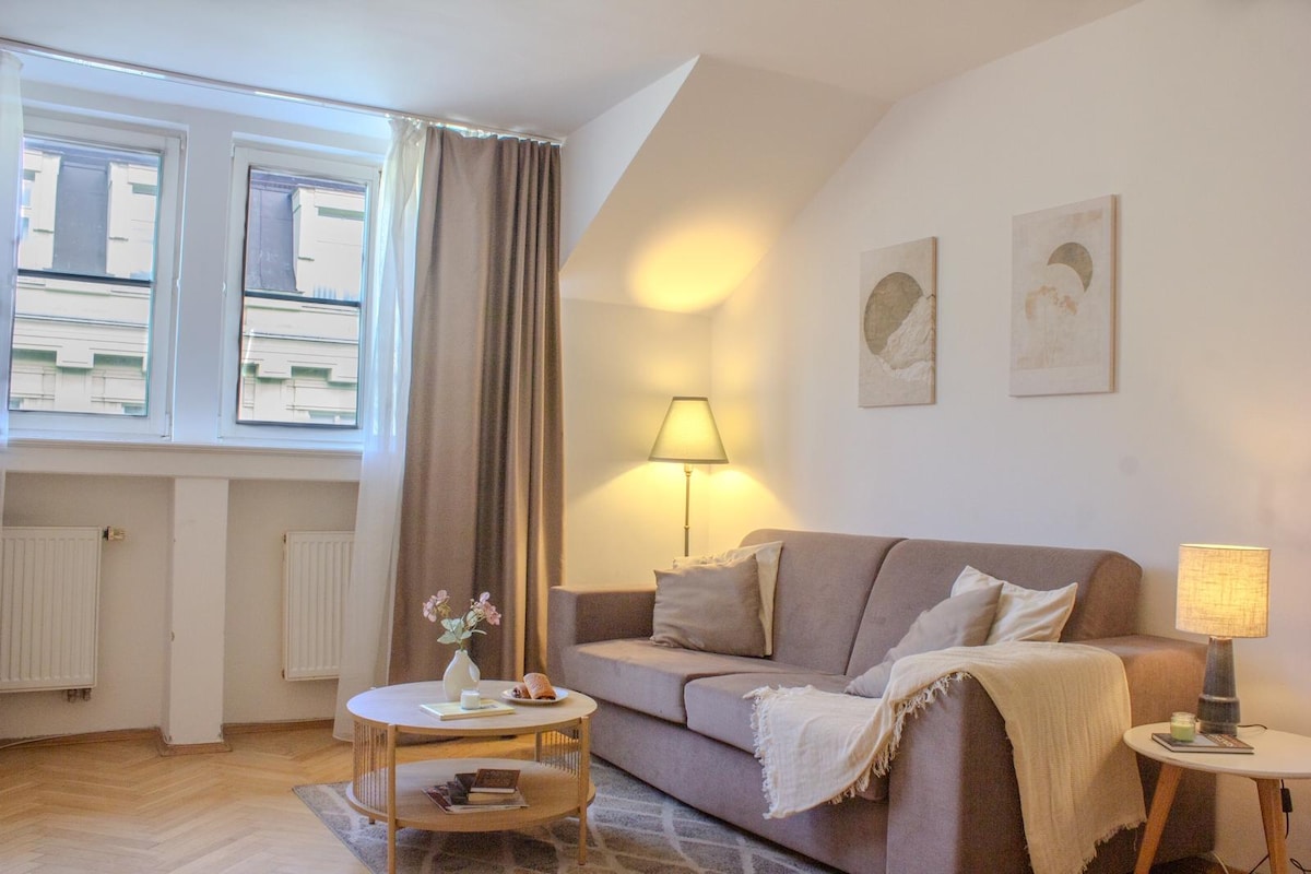 A cozy living room is illuminated by soft lighting, featuring a comfortable sofa with decorative pillows. A round coffee table is placed in front of the sofa, accompanied by a small vase of flowers. Two windows allow natural light to fill the space, complemented by light-colored curtains.
