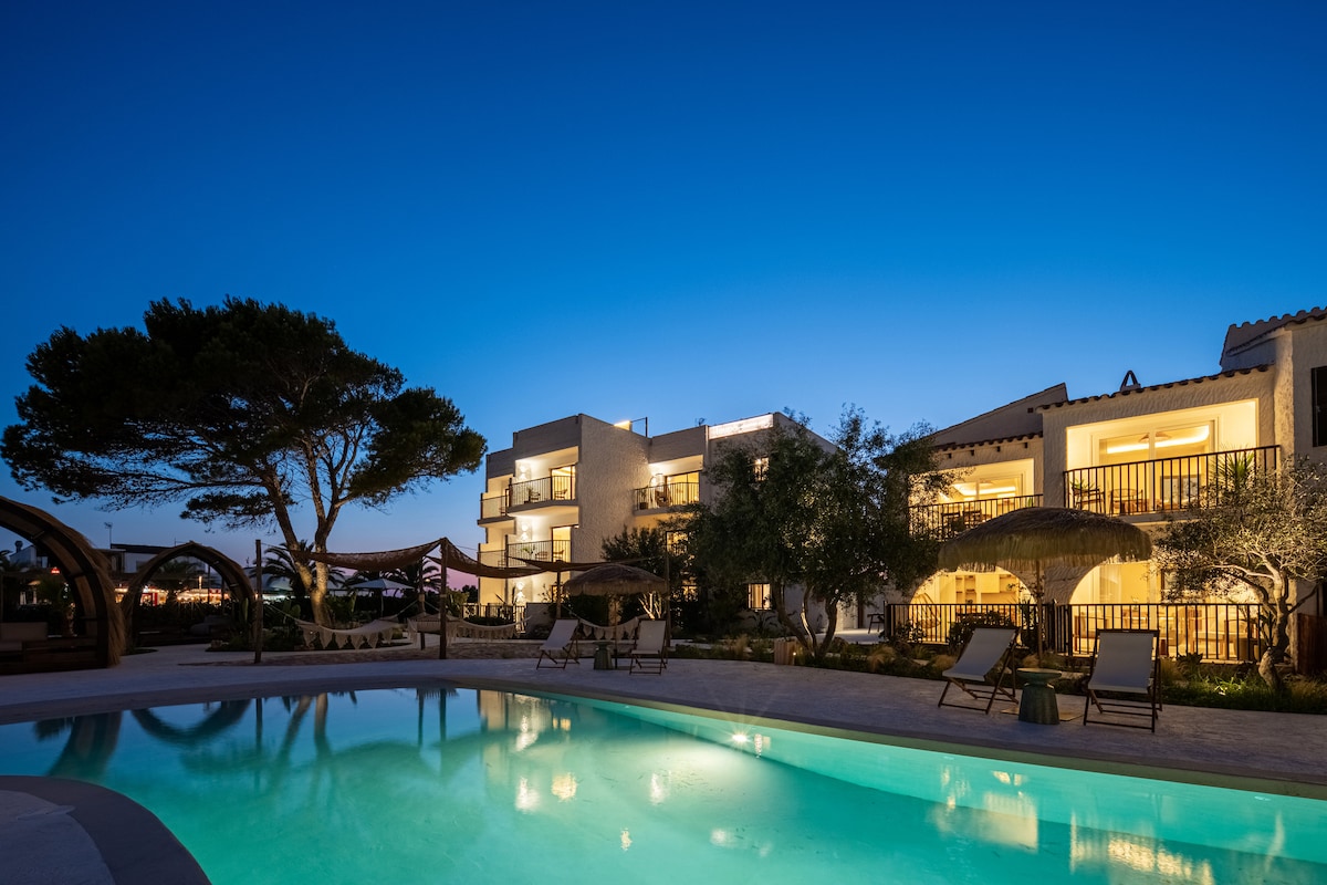 The tranquil pool area is illuminated under a deep blue sky, with modern buildings that feature balconies overlooking the water. Comfortable loungers are positioned around the pool, shaded by large umbrellas and accompanied by lush greenery, creating a serene outdoor environment.