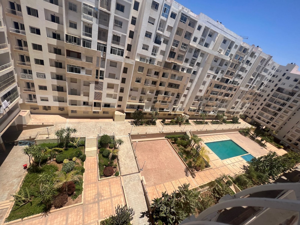 A view from an upper floor showcases a well-maintained outdoor area, featuring a swimming pool surrounded by landscaped gardens. Several pathways lead through the greenery, while neighboring apartment buildings create a sense of community in the serene environment.