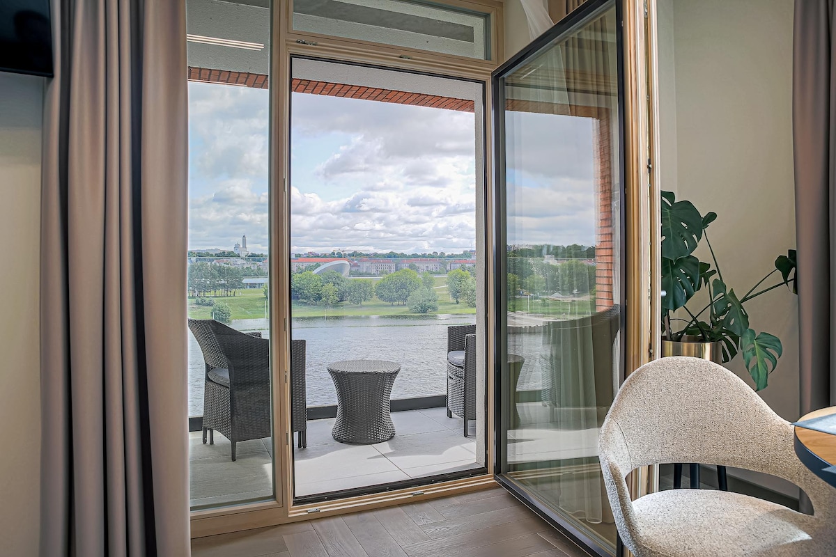 A sliding glass door allows access to a private balcony with views of a serene waterway and lush greenery. Comfortable outdoor seating is arranged beside a round table. Soft drapery frames the door, enhancing natural light that fills the indoor space.