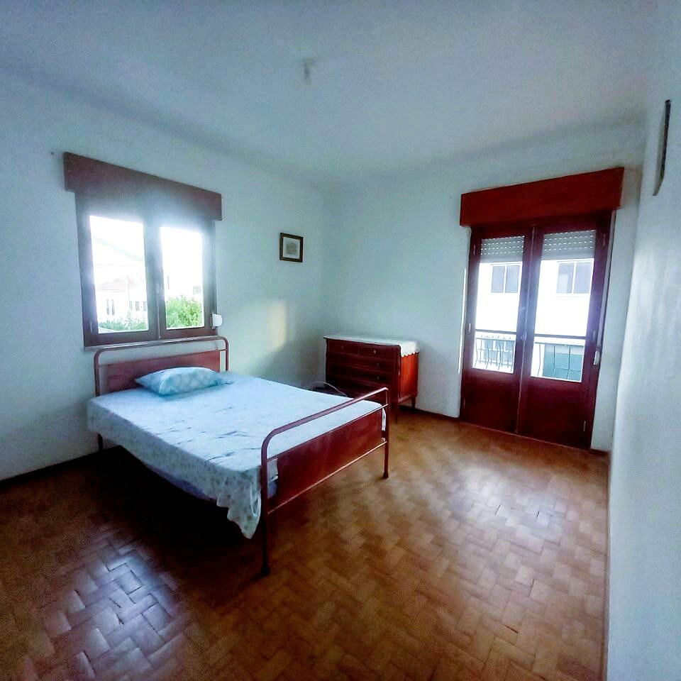 A spacious bedroom is presented with light wood flooring and white walls. Two large windows allow natural light to fill the room. A bed frame is positioned against one wall with a simple mattress, while a dresser stands nearby, completing the minimalistic design.