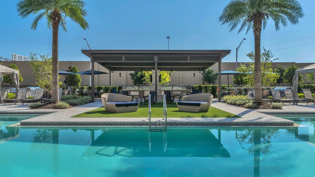 A swimming pool is surrounded by tall palm trees and comfortable lounge seating. Canopies provide shaded areas, while vibrant greenery enhances the inviting atmosphere. The pool reflects the clear blue sky, creating a serene setting for relaxation.