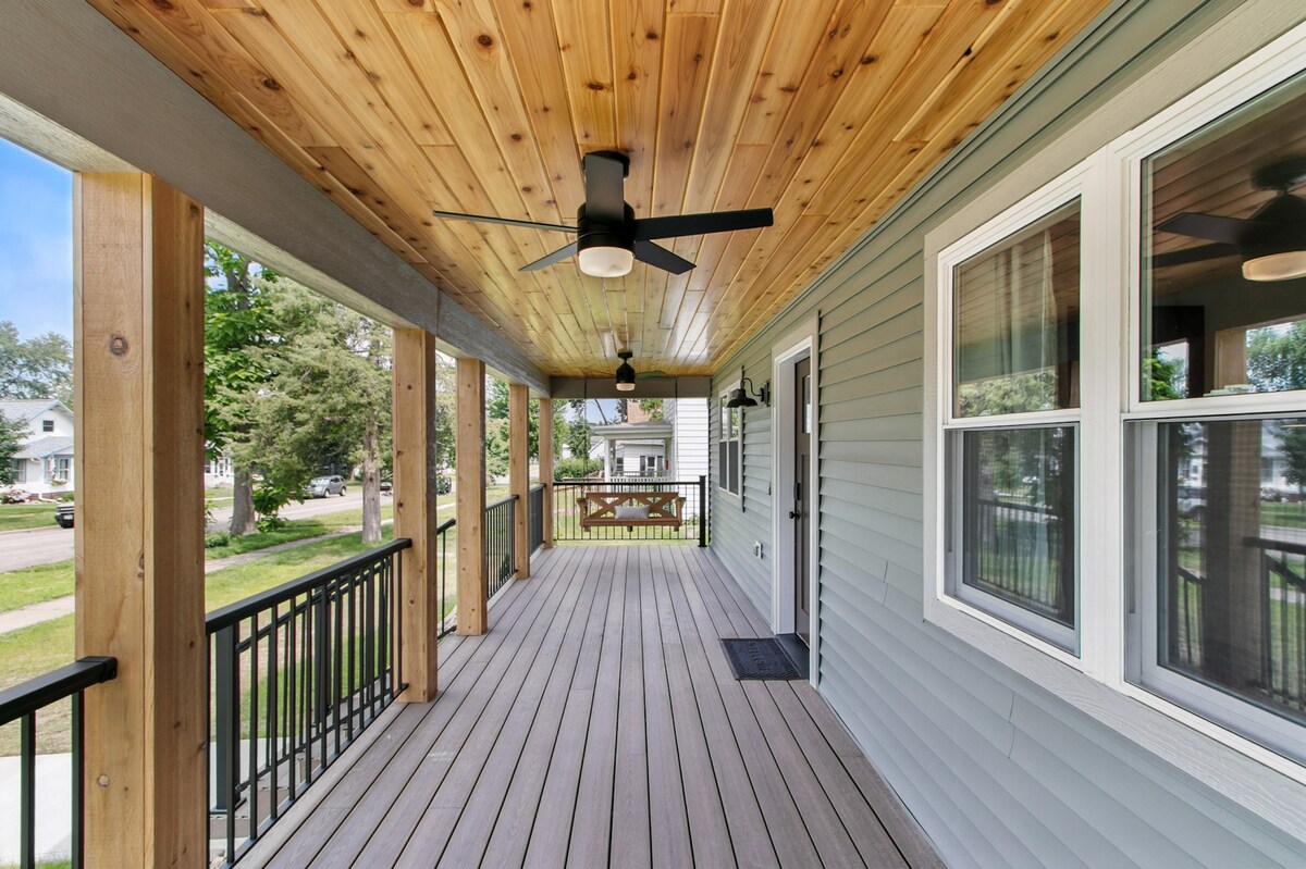 A spacious outdoor deck is shown, featuring wooden flooring and a ceiling with natural wood paneling. Ceiling fans are visible, providing airflow. A rail with black metal accents borders the deck, and large windows offer views of the surrounding neighborhood.