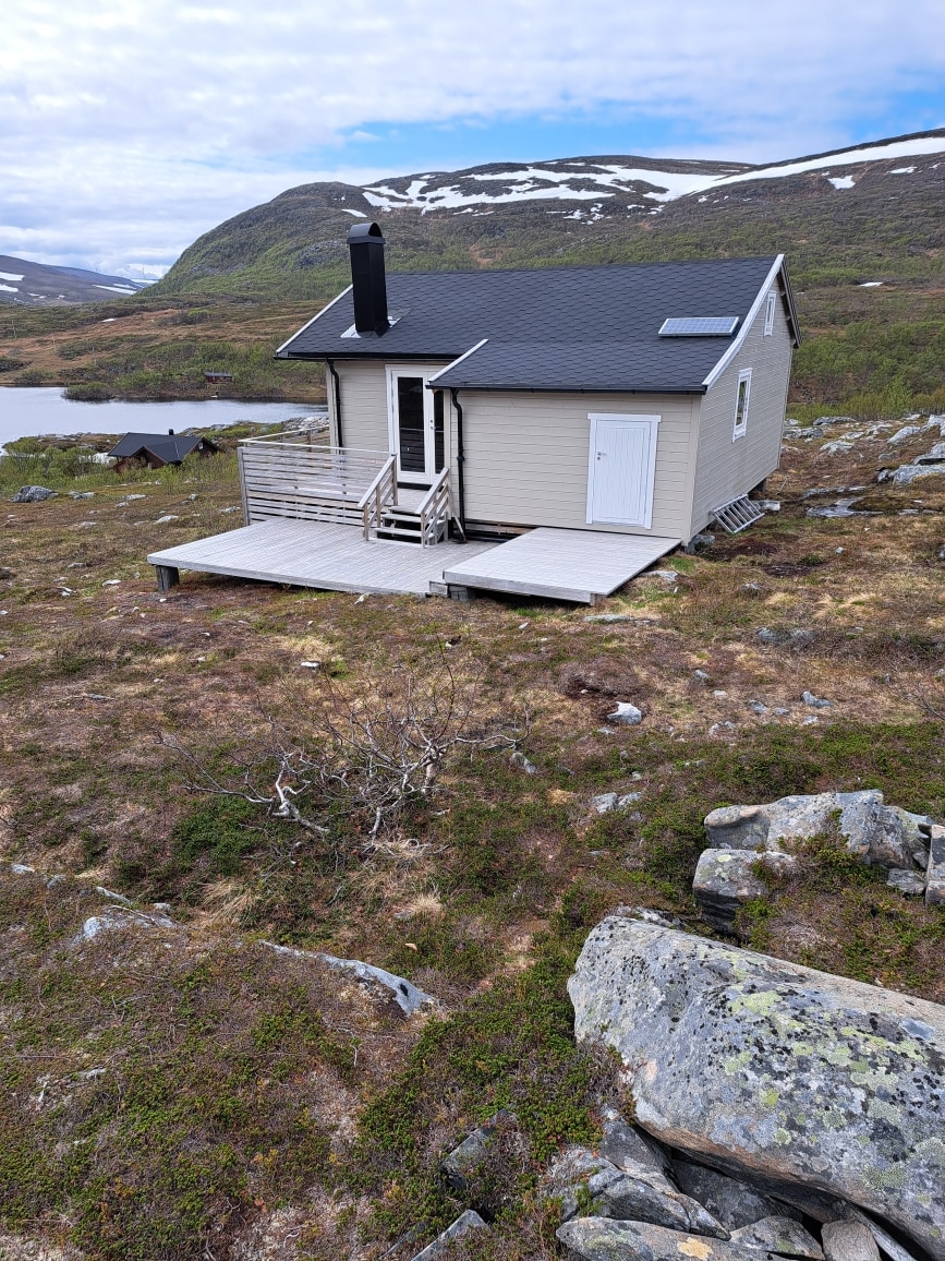 A quaint cabin is situated on a grassy hillside, surrounded by rocky terrain and a serene mountain backdrop. The cabin features a small deck with steps leading to the entrance. Solar panels are visible on the rooftop, providing a sustainable energy source.