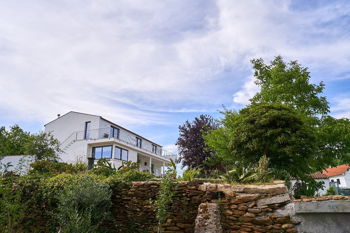 A contemporary house is seen from the garden, featuring a multi-level design with large windows. Lush greenery surrounds the exterior, complemented by a stone wall that adds natural texture. Soft clouds drift across a blue sky, hinting at a serene atmosphere.