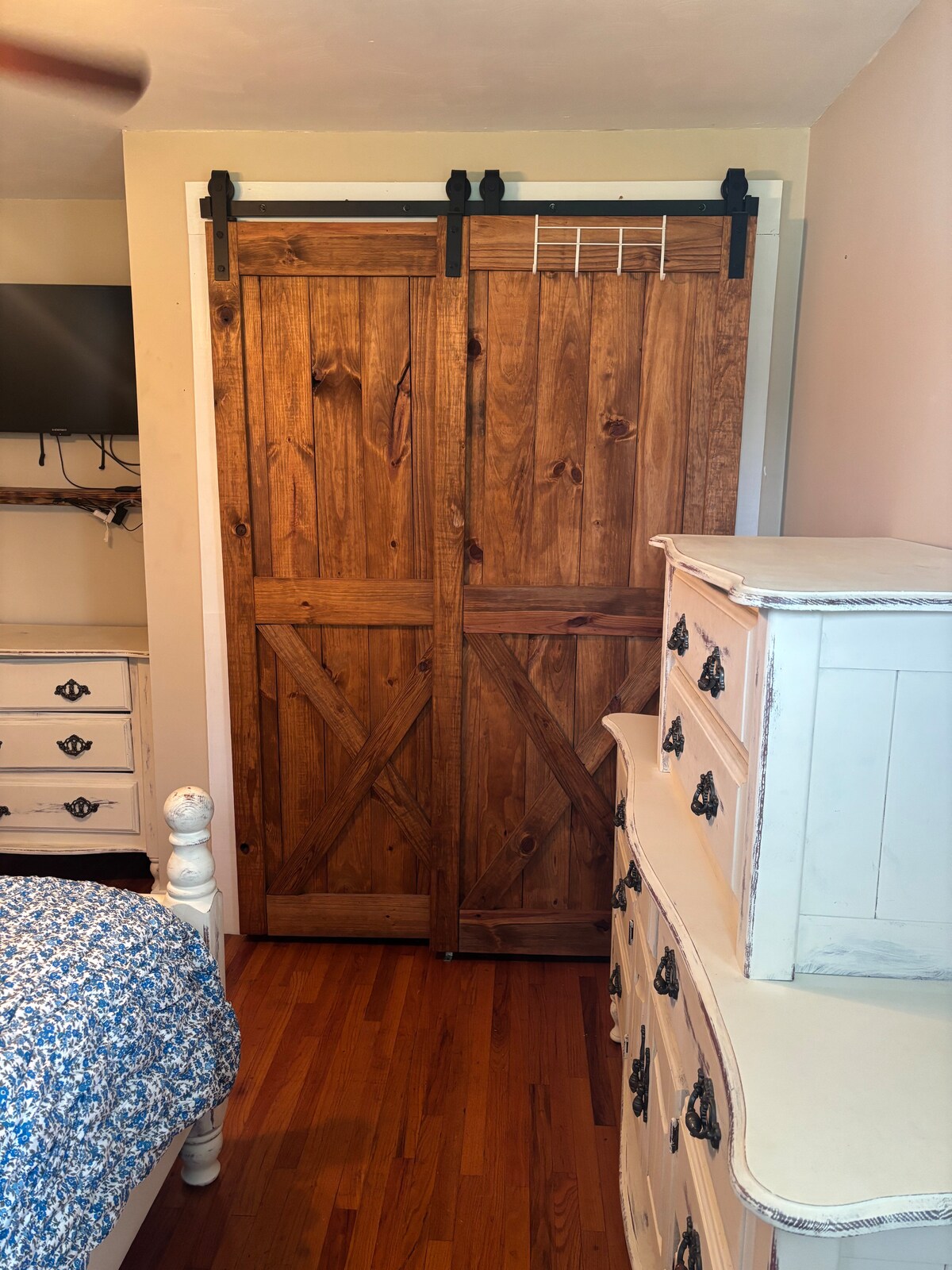 A rustic wooden sliding barn door is featured, leading to a storage area, complemented by a light finish dresser nearby. The hardwood floor adds warmth to the space, with a cozy bed adorned in floral patterned bedding visible in the foreground.