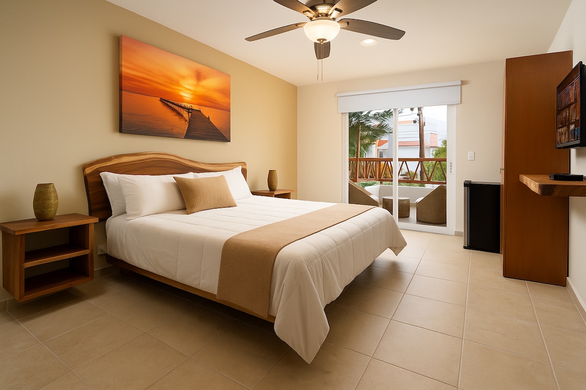 A spacious bedroom features a king-size bed with neutral-toned bedding. Two wooden nightstands with decorative vases are positioned beside the bed. A large window offers views of a terrace, while a mounted TV and a small fridge are present within the room.