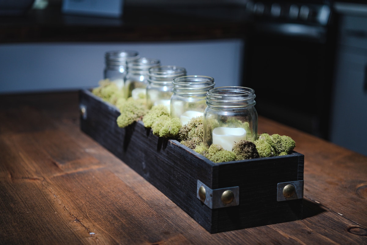 A long wooden centerpiece holds several glass jars filled with candles, surrounded by a bed of textured moss. The arrangement is set on a rustic wooden table, with soft ambient light enhancing the natural elements.