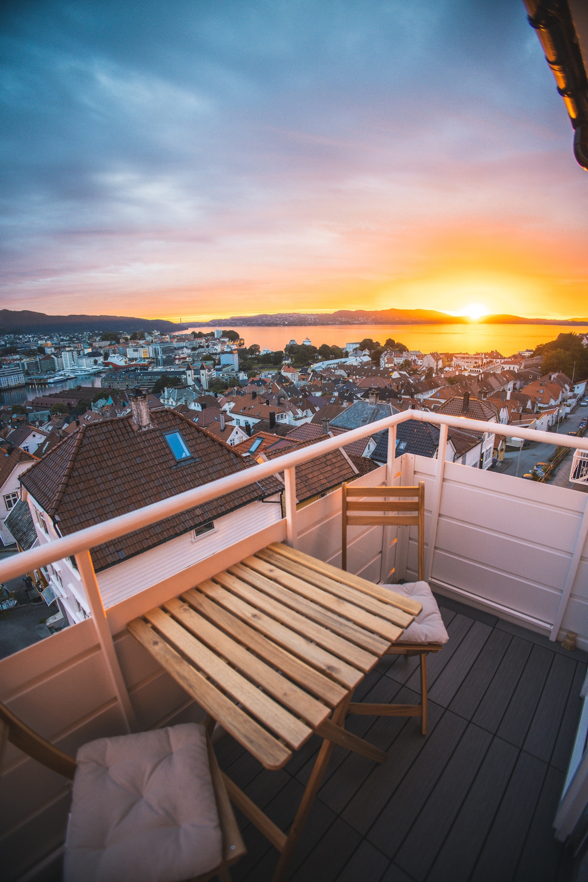 A balcony is furnished with a wooden table and two chairs, offering a view of the sunset over the water. The surrounding rooftops and distant hills create a scenic backdrop, while the sky displays warm hues of orange and purple.