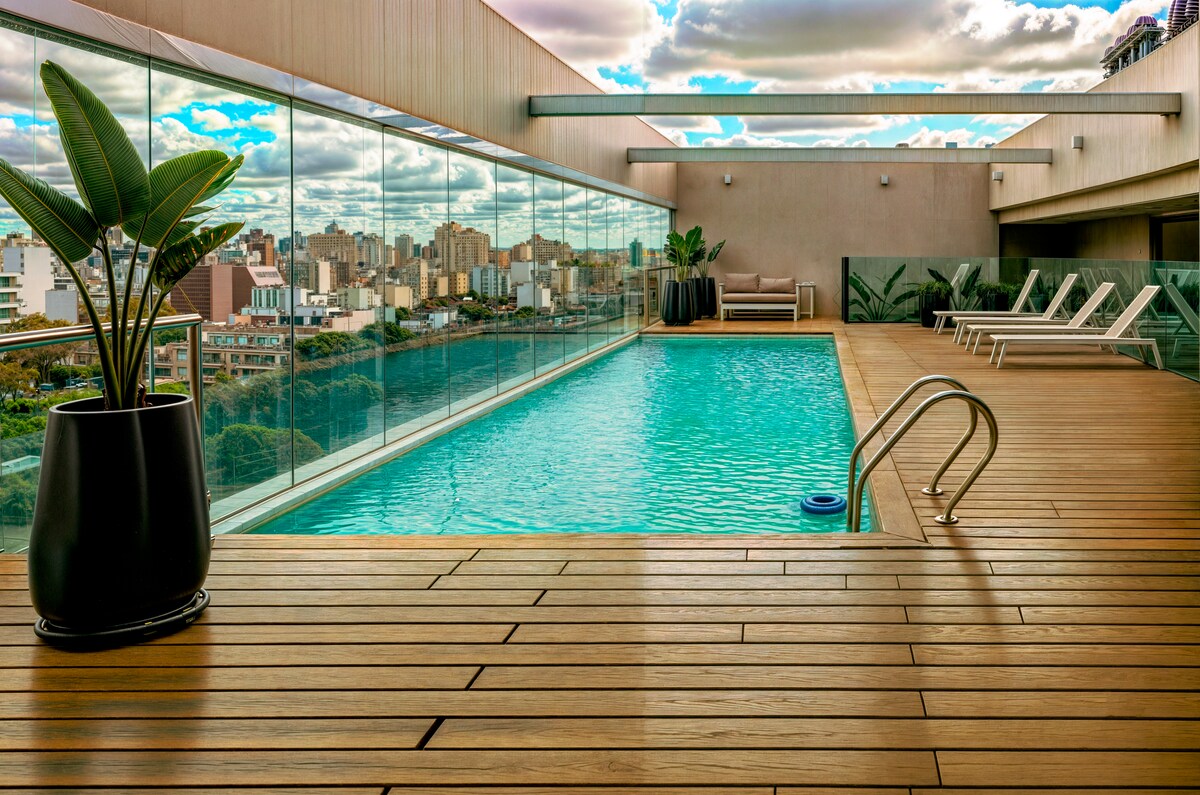 An elegant outdoor pool area features a clear blue pool surrounded by wooden decking. Comfortable lounge chairs are positioned beside the pool, with modern planters adding greenery. A panoramic view of the city skyline is visible through the large glass barriers.
