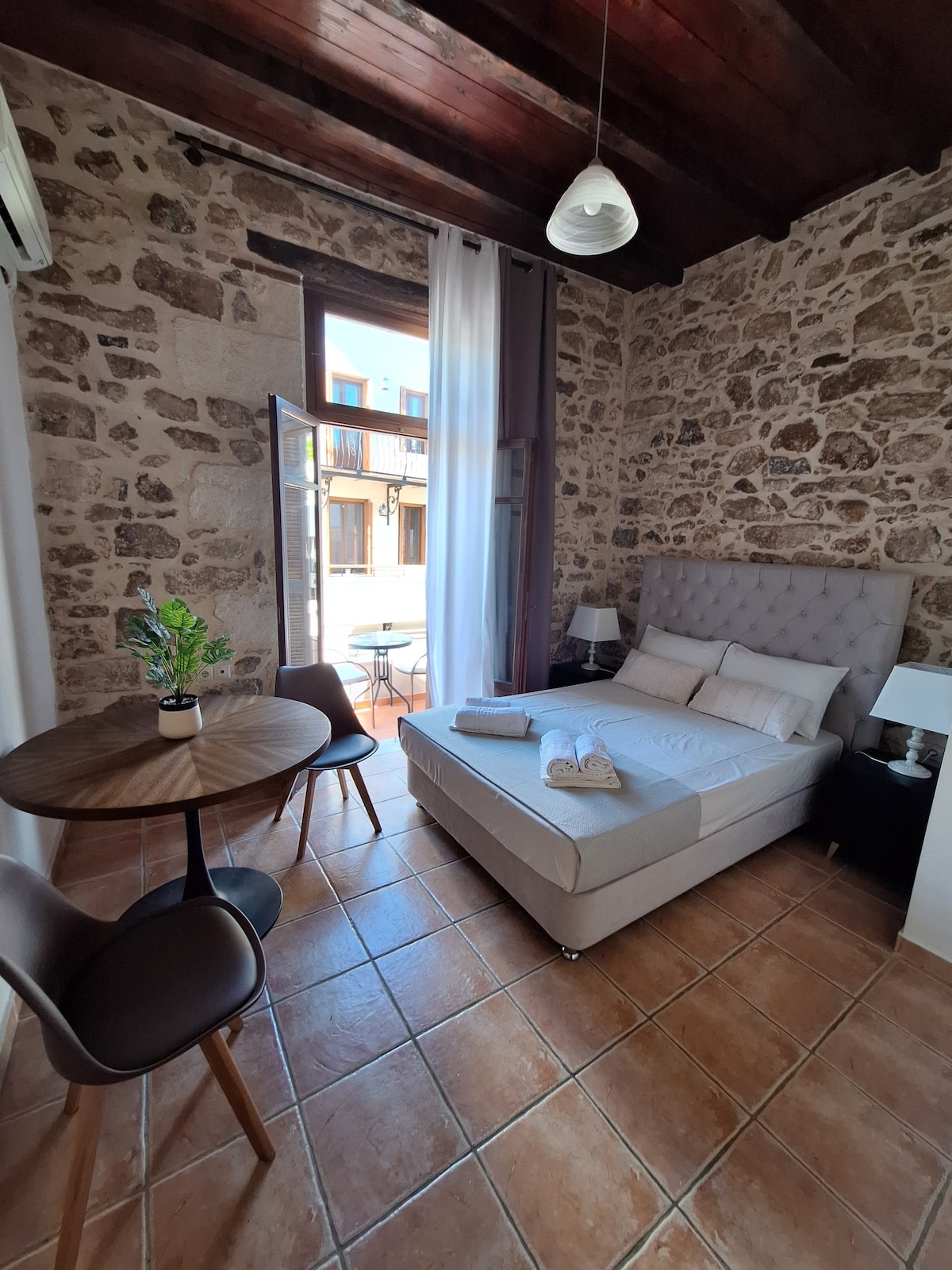 A cozy room features a spacious bed with light-colored linens, complemented by a round dining table with two chairs. Exposed stone walls and warm tile flooring add character, while a large window illuminates the space and offers a view of the outdoor area.