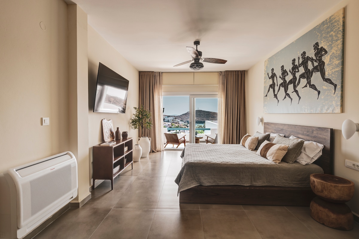 A spacious bedroom features a large bed with layered linens and decorative cushions. A television is mounted on the wall, and a stylish side table is placed nearby. Artwork depicting figures in motion hangs above the bed, with sliding glass doors leading to a balcony offering sea views.