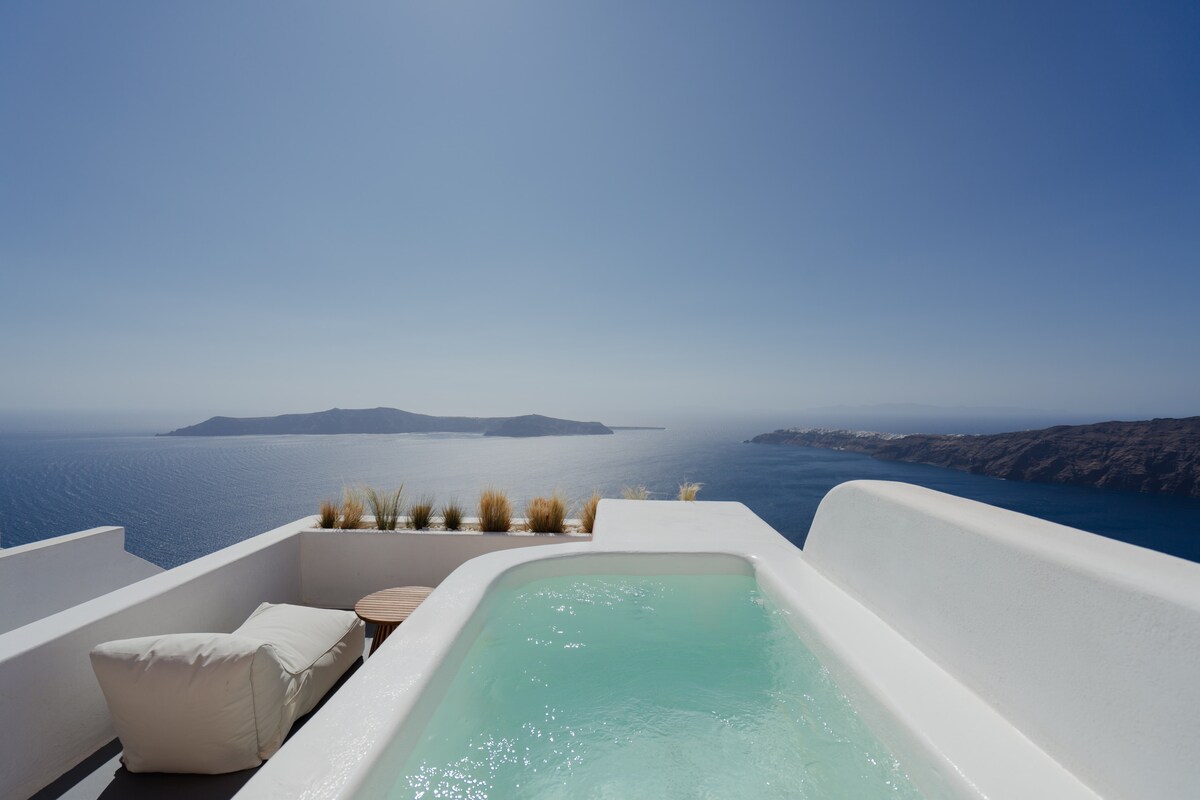 An open-air hot tub filled with clear water is positioned on a spacious terrace. The view extends over the deep blue Aegean Sea, displaying distant islands against a bright blue sky. Soft greenery lines the terrace, complementing the serene atmosphere.