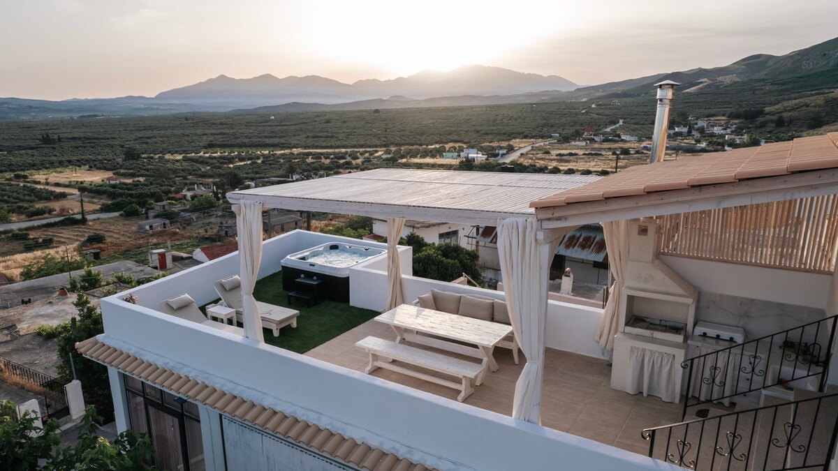 An elevated terrace features a private jacuzzi and outdoor seating area, surrounded by sheer curtains. Scenic views of lush valleys and mountains are visible in the distance, with a barbecue grill positioned nearby, enhancing the outdoor experience.