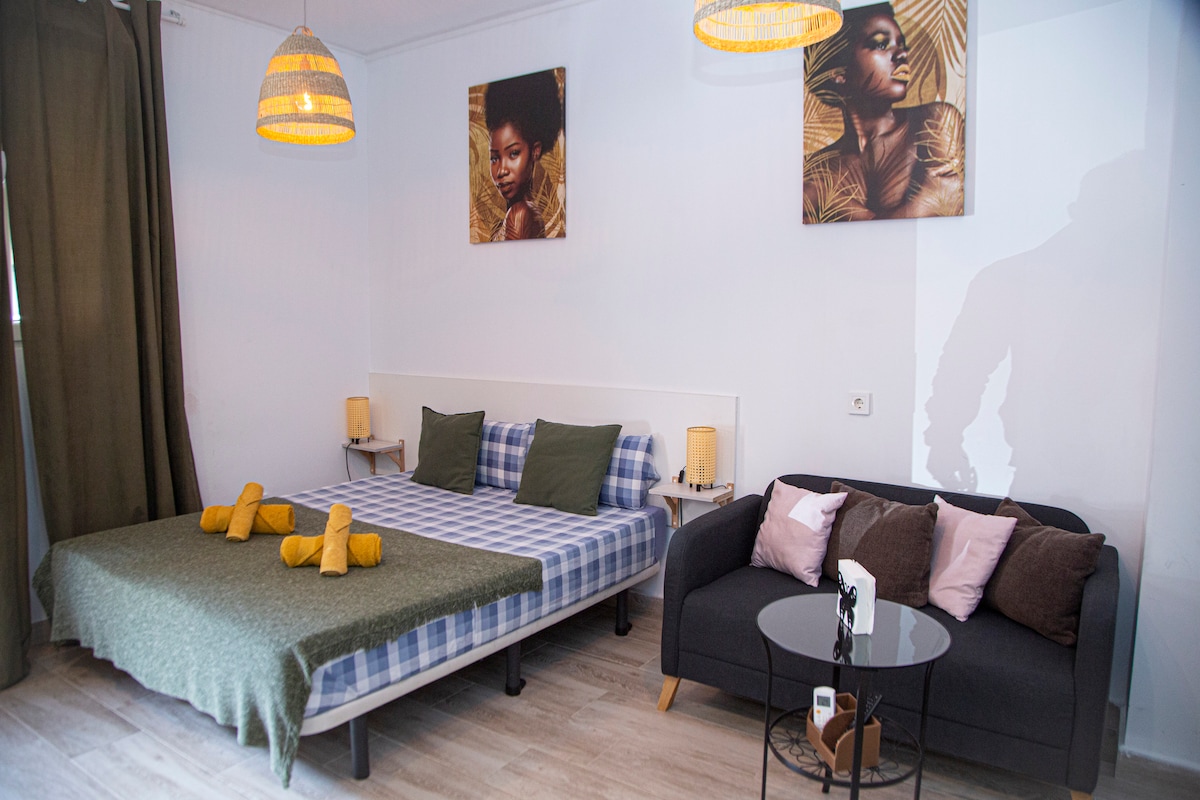 A spacious sleeping area features a comfortable bed dressed with a patterned blanket and cushions. Two side lamps offer soft lighting, while artwork adorns the walls. A cozy sofa sits opposite the bed, accompanied by a small circular table, enhancing the inviting layout.