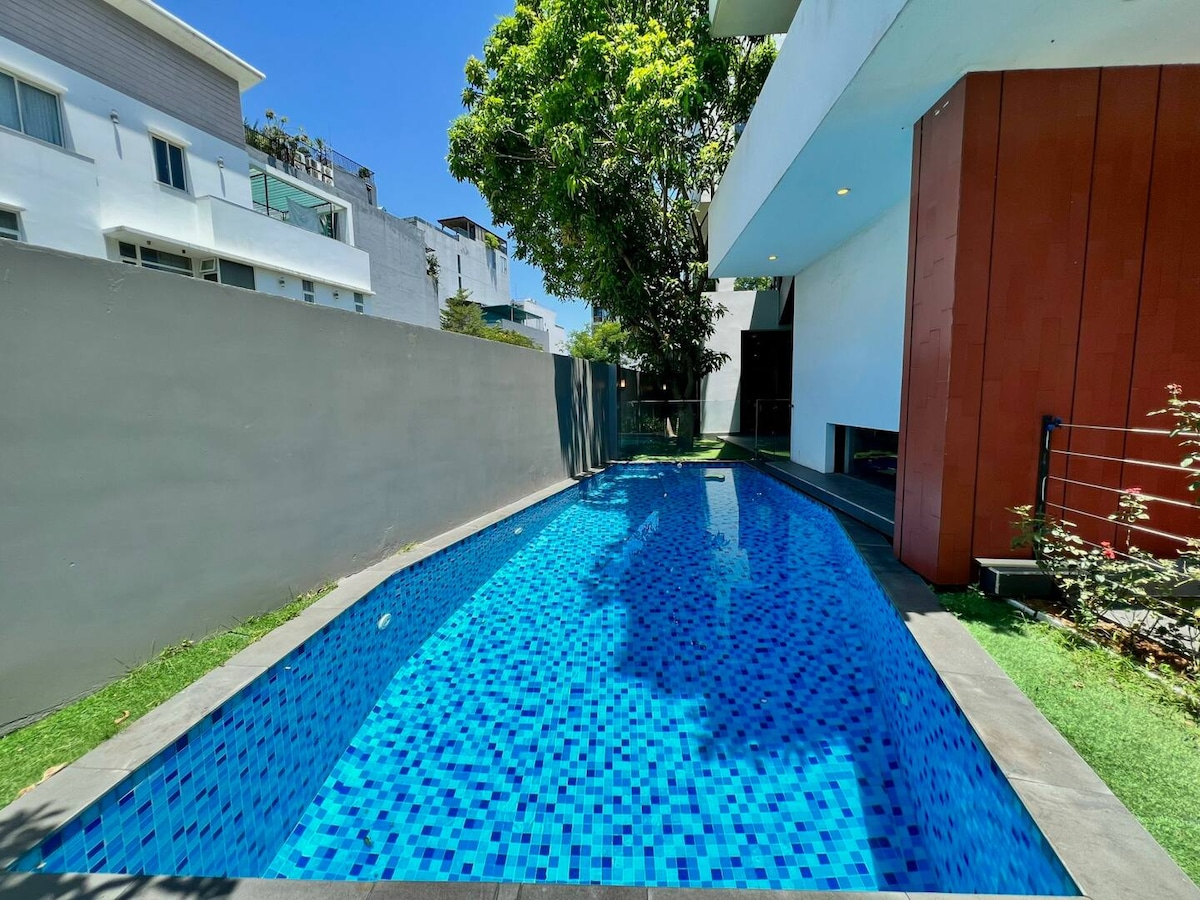 A private swimming pool is framed by a modern villa, featuring bright blue tiles that reflect sunlight. Lush greenery surrounds the pool area, enhancing the sense of outdoor relaxation. The clear sky above complements the inviting atmosphere of the space.