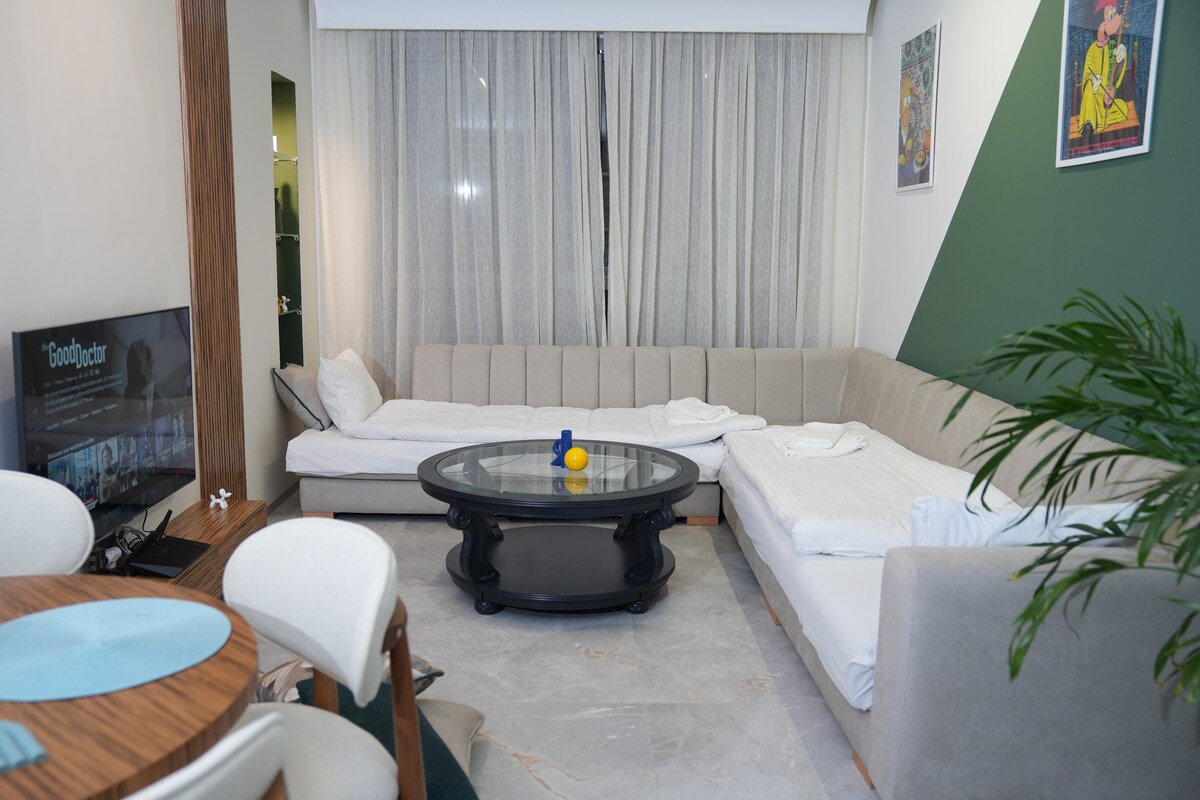 ამ თანამედროვე და ელეგანტური მისაღები ოთახის სივრცეში თავმოყრილია დასასვენებელი ზონა და საუზმისთვის განკუთვნილი მაგიდა. ქოთნებში მოთავსებული მცენარეები და ნათელი ფერების პრინტები სივრცეს ინდივიდუალურობასა და ხიბლს სძენს.