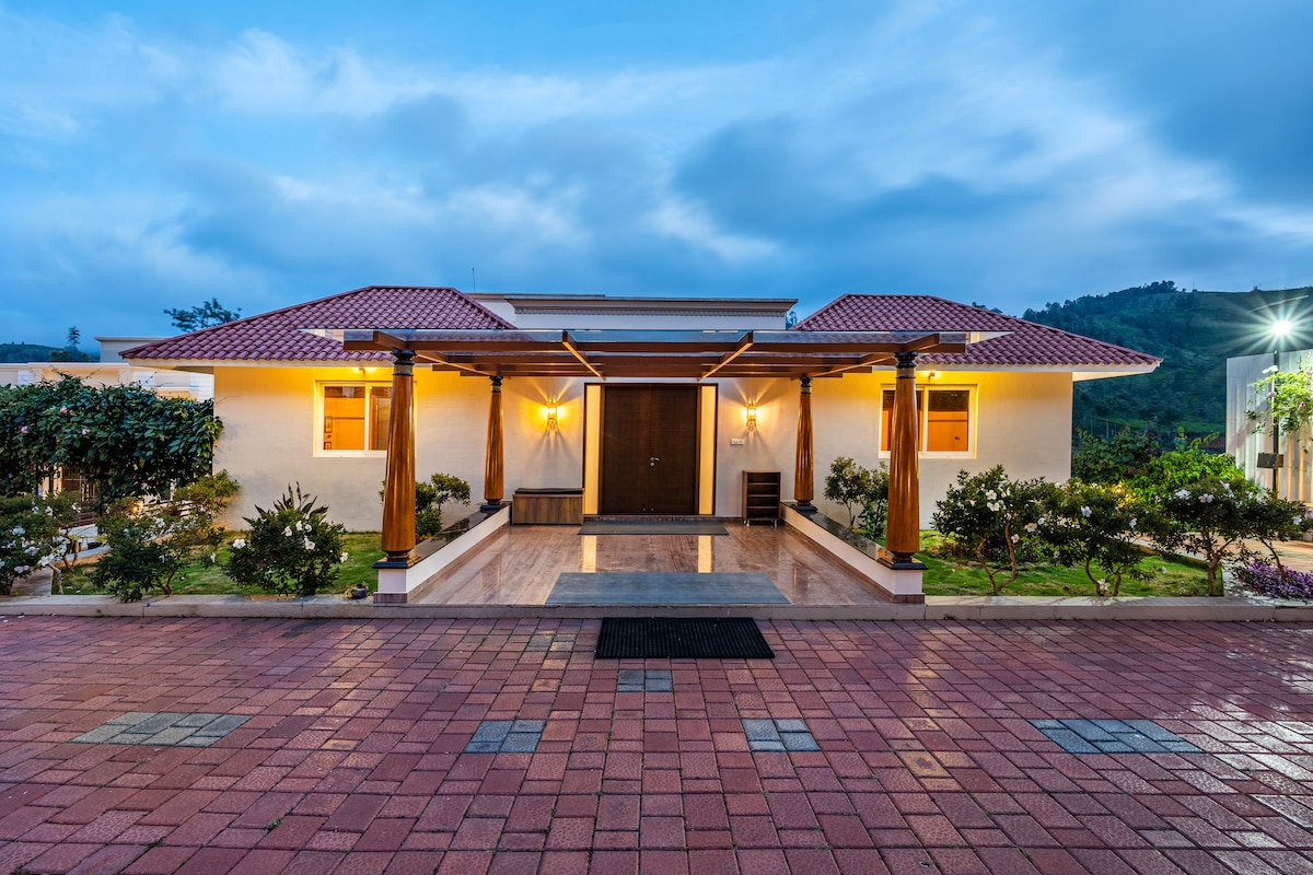 A modern villa façade is displayed, featuring a spacious entrance with polished stone flooring. The structure is flanked by well-maintained greenery and framed by wooden pillars, while two symmetrical roofs with red tiles provide a welcoming touch. Soft lighting enhances the entrance appearance.