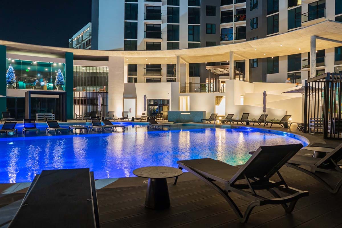 A modern outdoor pool area is illuminated at night, showcasing a large semi-olympic pool surrounded by lounge chairs and umbrellas. The sleek building architecture contrasts with the tranquil water, offering a relaxing atmosphere.