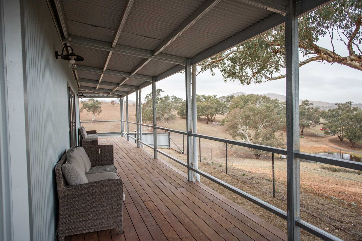 A wide veranda features wooden flooring and railings, offering a serene view of surrounding hills and tree lines. Two comfortable wicker chairs are positioned along the veranda, creating a space for relaxation and connection with nature.
