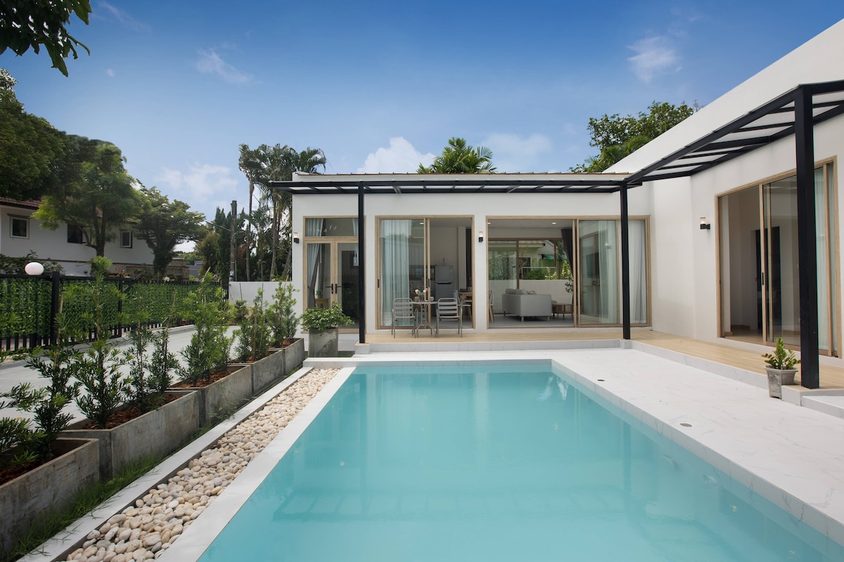 A private villa is showcased with a serene pool in the foreground. The entrance is characterized by large glass doors opening to a bright interior, framed by modern architecture. Lush greenery surrounds the property, blending nature with a minimalist design.