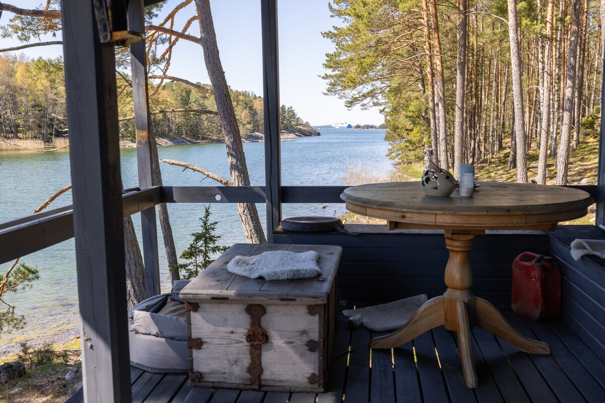 A wooden table with a round top is positioned in a secluded outdoor space. A rustic chest sits beside it, and soft textiles are laid on the seating area. Views of calm water and distant islands are framed by tall trees, creating a serene atmosphere.