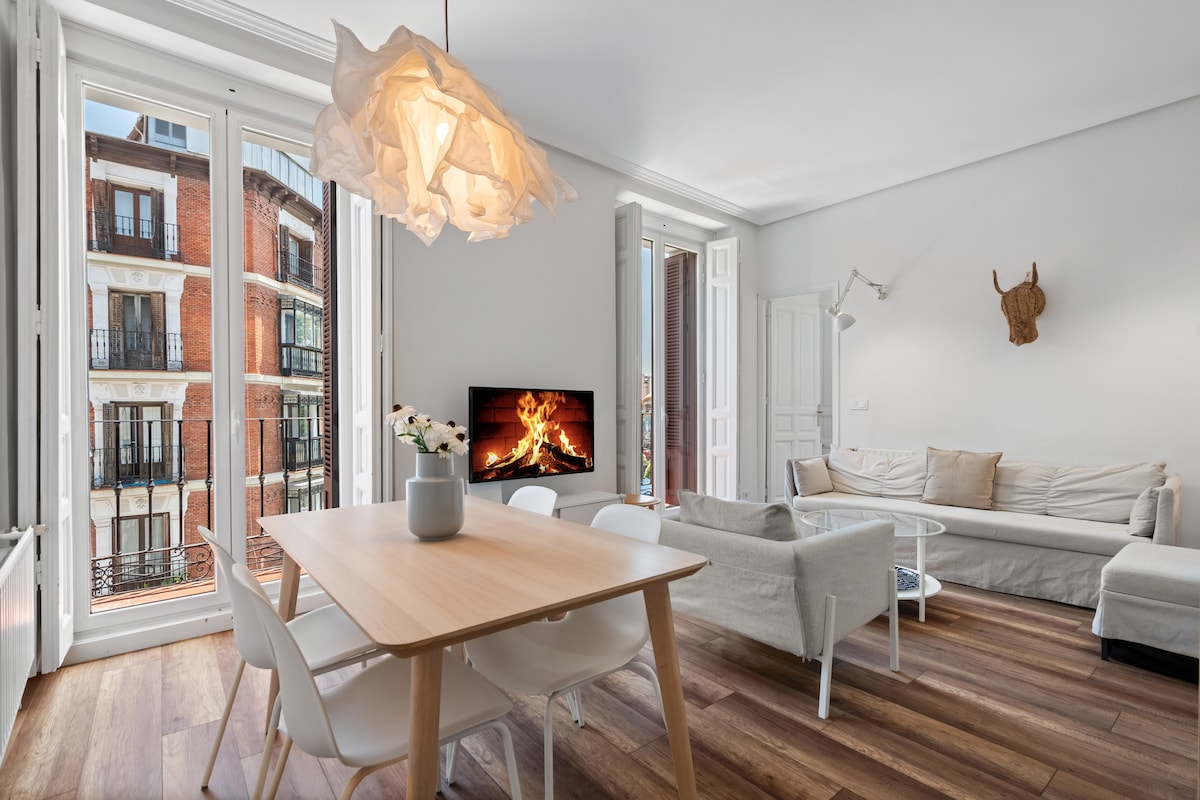 A bright living area features large windows that provide views of the street. A wooden dining table is surrounded by white chairs, while a comfortable sofa is positioned nearby. A modern light fixture hangs from the ceiling, adding a touch of style to the space.