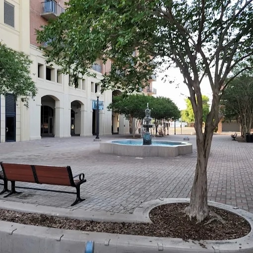 A serene plaza area features a central fountain surrounded by well-maintained landscaping. Several trees provide shade, while a single wooden bench offers seating. The brick-paved ground is organized and tidy, creating an inviting outdoor space.
