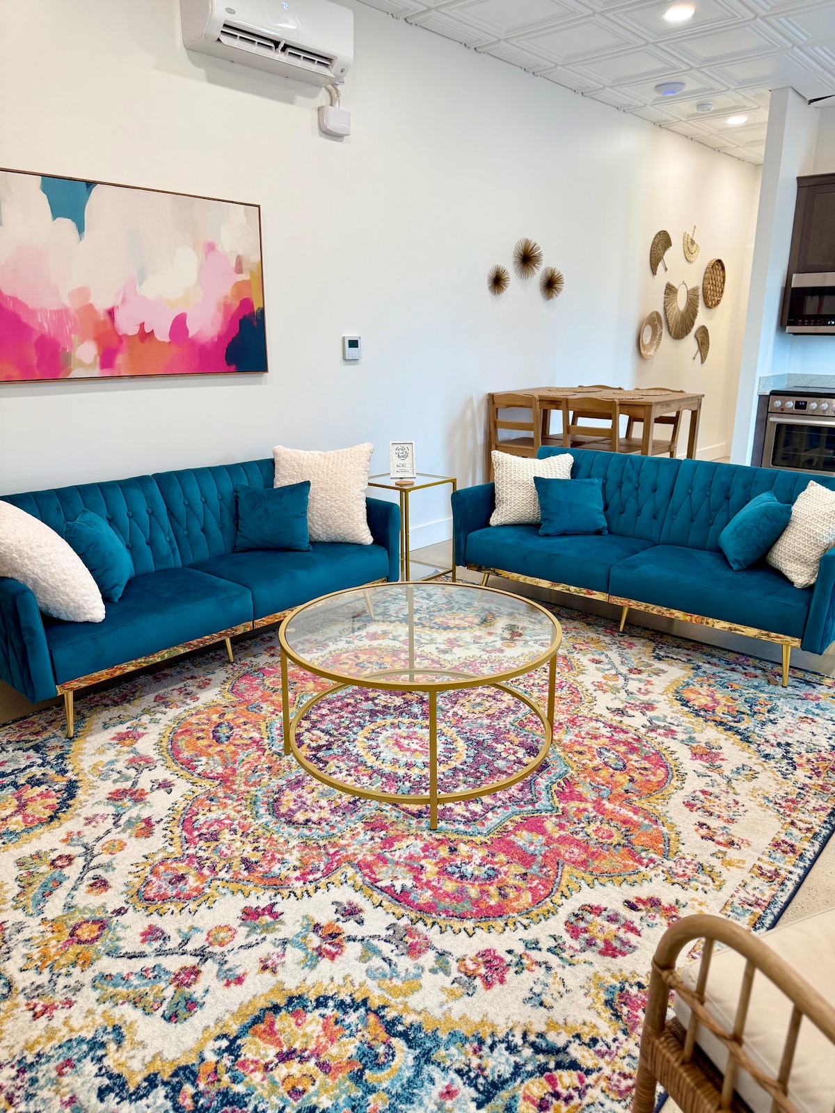 A bright living area features a pair of deep blue sofas adorned with decorative pillows, surrounding a circular coffee table. A colorful area rug with intricate patterns covers the floor, and a light artwork hangs on the wall, complemented by woven decorative elements.