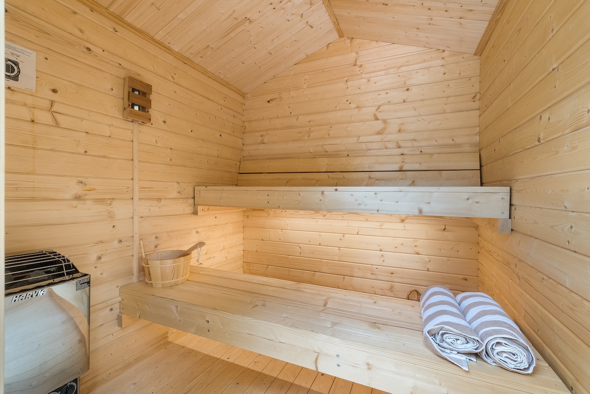 A serene sauna interior is showcased with smooth wooden paneling and two tiered benches. A traditional sauna stove is positioned to the left, accompanied by a wooden bucket and fresh towels neatly rolled on the right.