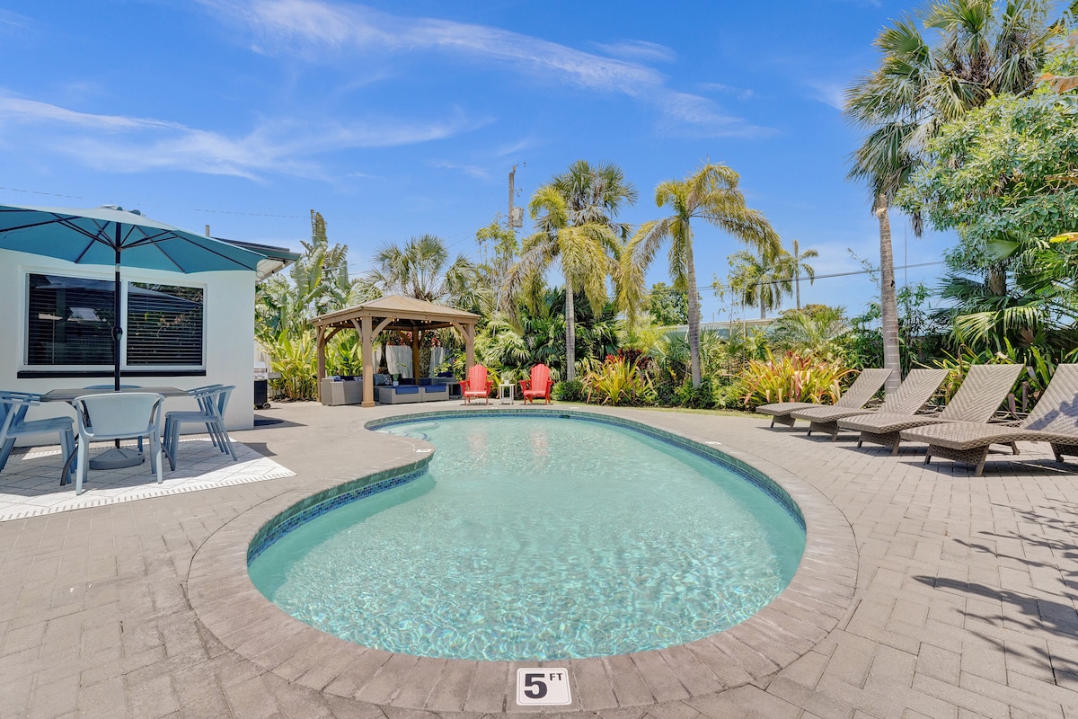 A private pool area features a heated, sparkling pool surrounded by tropical landscaping. Comfortable sun loungers are positioned nearby, and a shaded outdoor living room is visible under a gazebo, inviting relaxation in a serene environment.