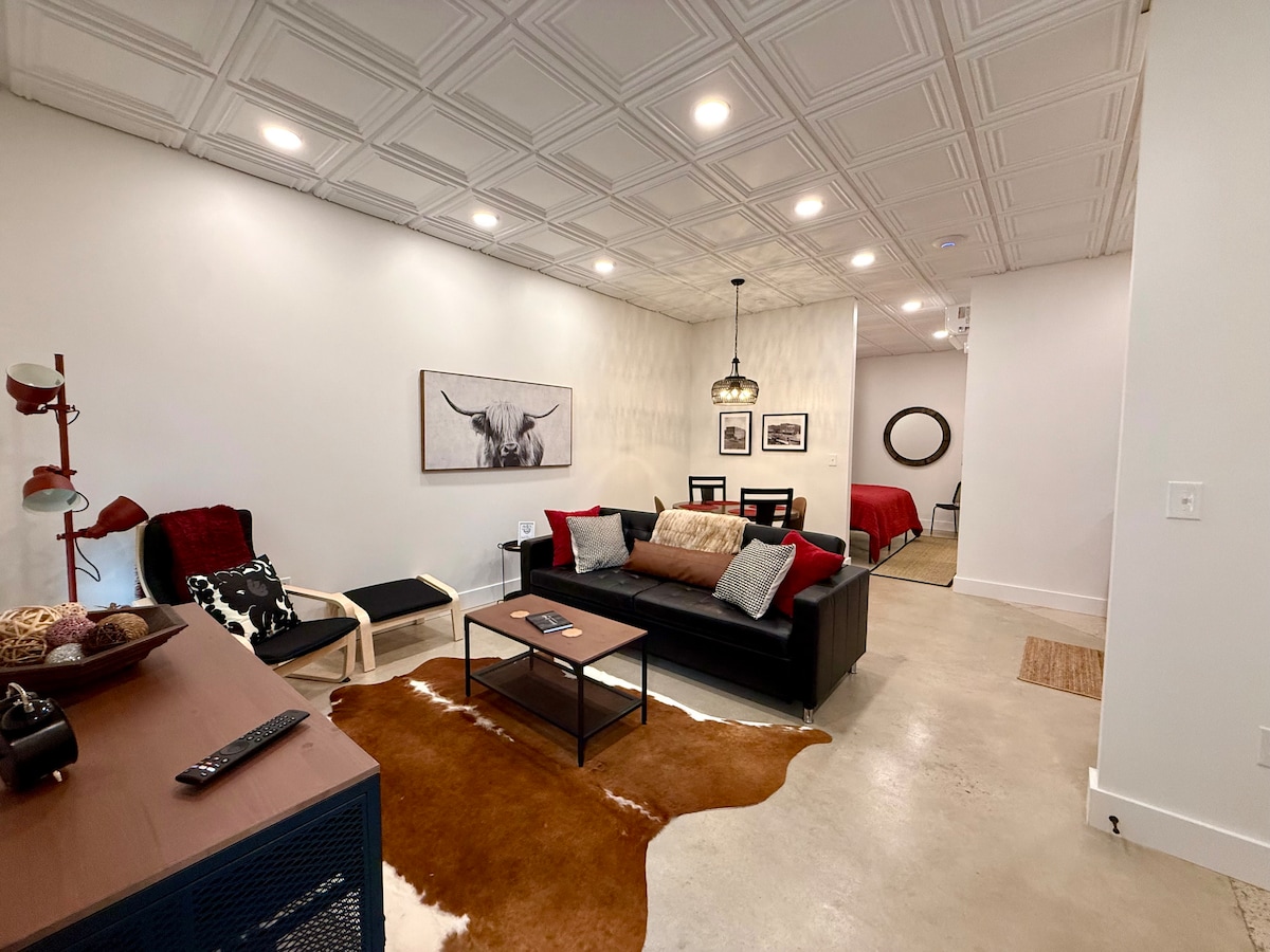 The living area presents a modern design, featuring a dark sofa adorned with plush cushions. A coffee table sits on a patterned rug, and a floor lamp adds soft lighting. A wall-mounted art piece and a decorative chair enhance the industrial farmhouse aesthetic.